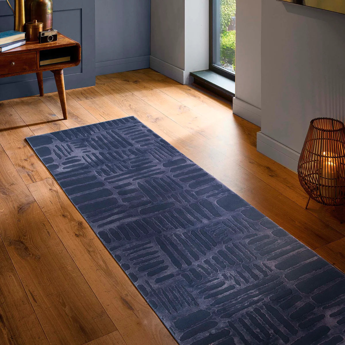 A long, dark blue non-slip rug rests on a wooden floor beside a window, with a lamp and wooden desk nearby in a modern room.