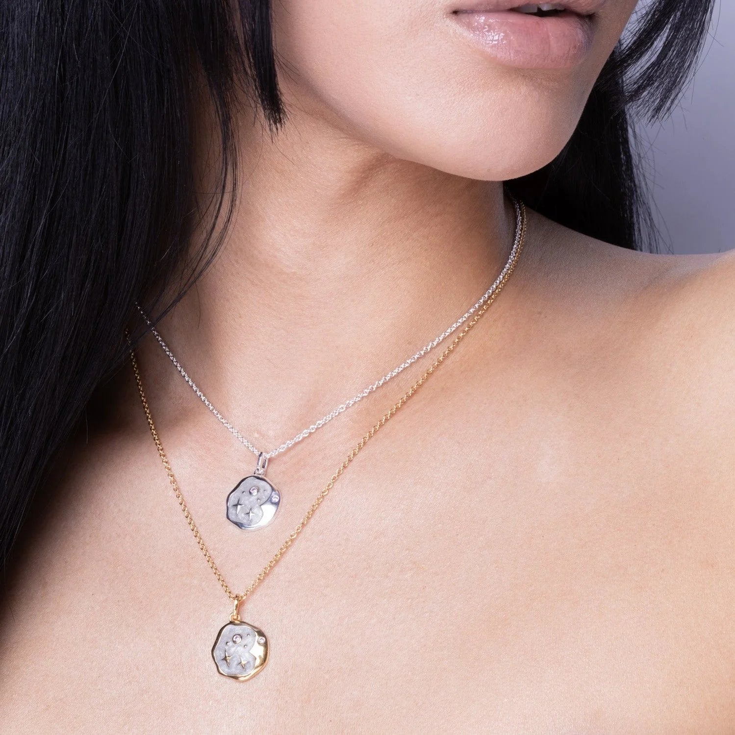 A person wearing two necklaces, one gold and one silver, each with a hexagonal pendant adorned with a clear gemstone—a striking pair inspired by celestial jewellery.