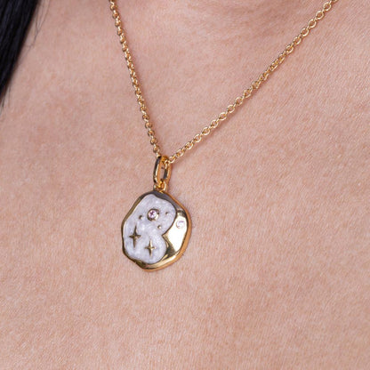 A gold celestial necklace featuring a textured, irregular-shaped pendant in white and gold tones, adorned with small embedded stones, elegantly drapes around the neck.