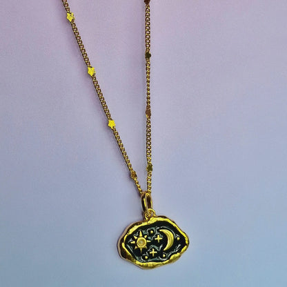 A black enamel and gold statement necklace with a rectangular pendant featuring celestial designs—sun, moon, and stars—on a dark background, elegantly displayed on a purple surface.