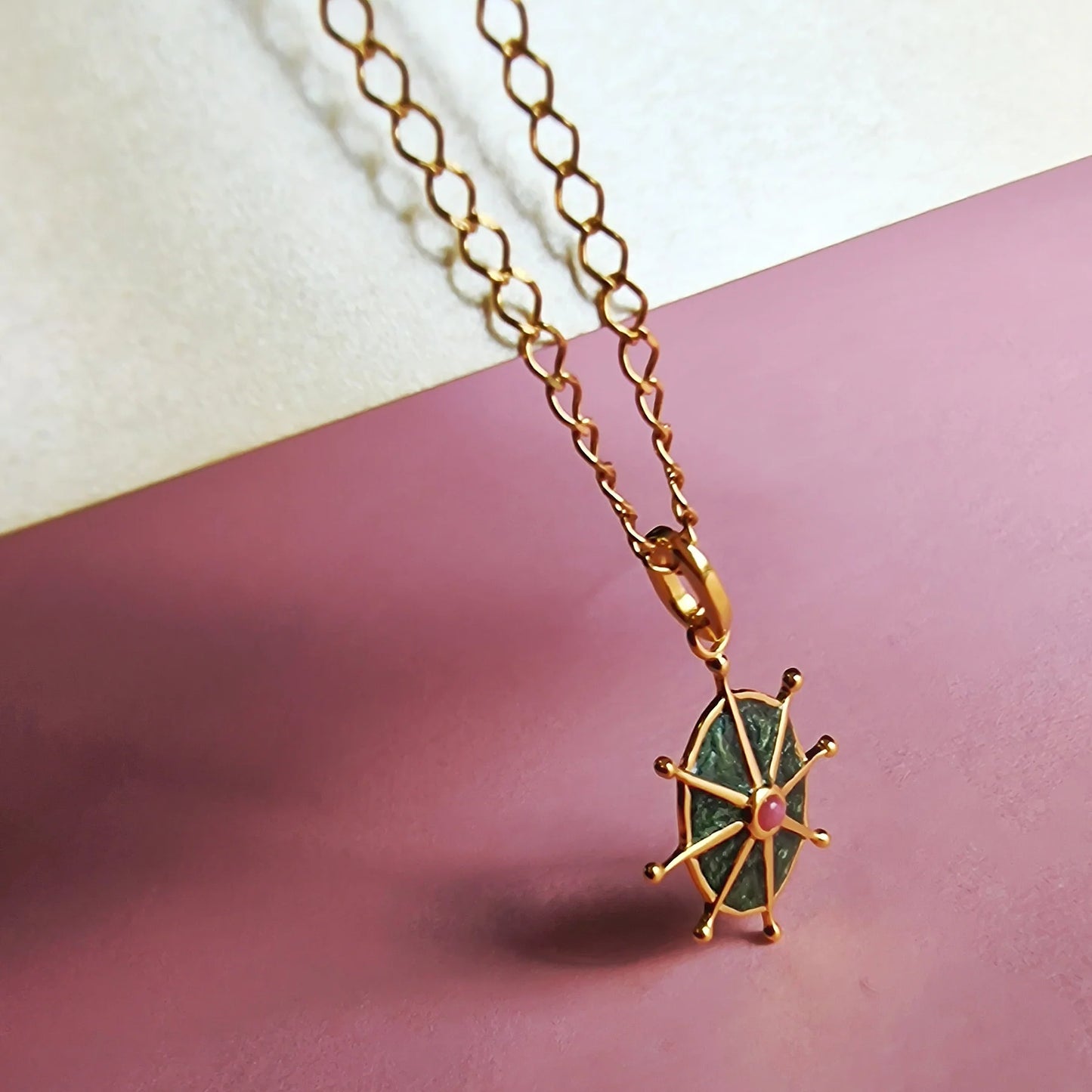 Gold chain necklace with a wheel-shaped pendant featuring a green enamel centre and a small pink stone, displayed on a pink and off-white background.