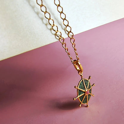 Gold chain necklace with a wheel-shaped pendant featuring a green enamel centre and a small pink stone, displayed on a pink and off-white background.