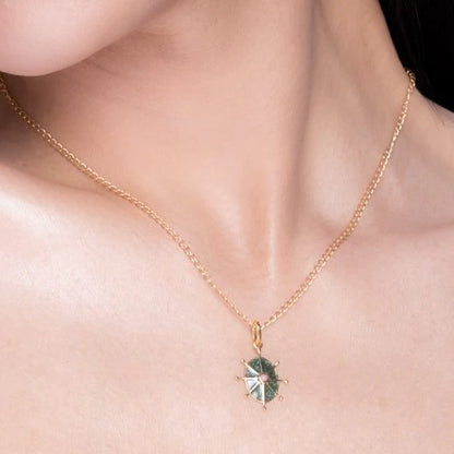 A gold chain necklace with a round green enamel pendant, showcasing green and white segments, is worn around a woman’s neck.