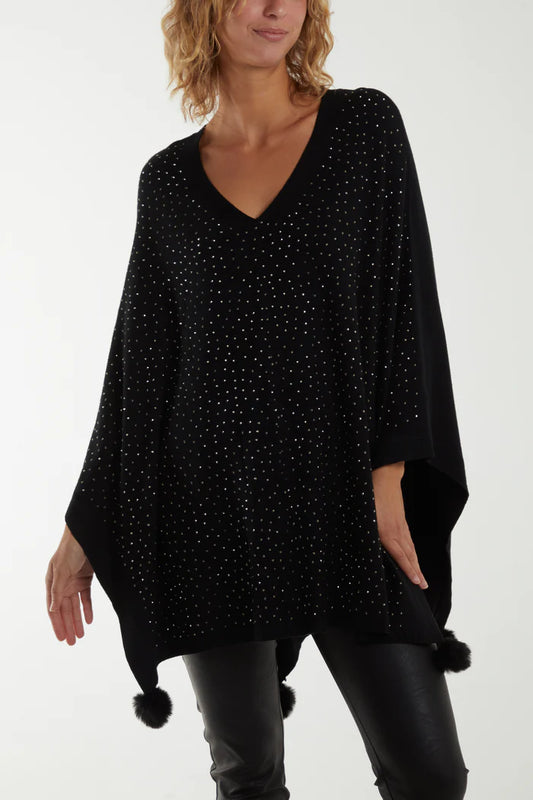 A woman wears a sparkly poncho with loose sleeves and black faux fur pom-poms at the corners, paired with black leather trousers—a stylish look ideal for fans of vegan fashion.