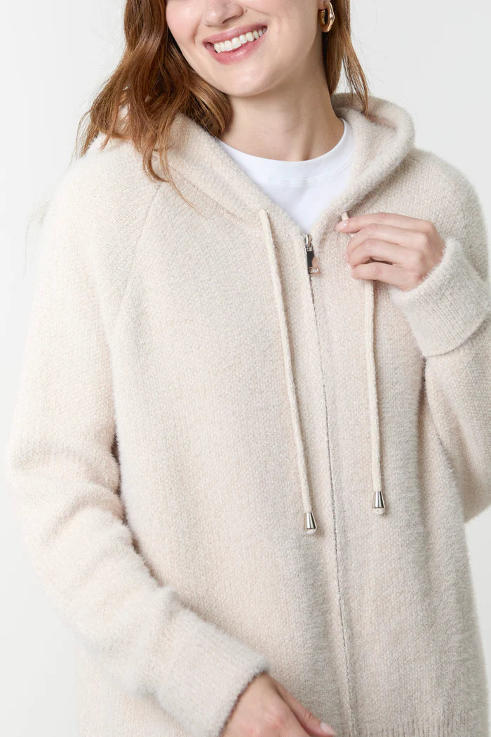 A person wearing a Super Soft Stone Hoodie, a cream-coloured vegan hoodie over a white shirt, holding the zip with one hand and smiling slightly.