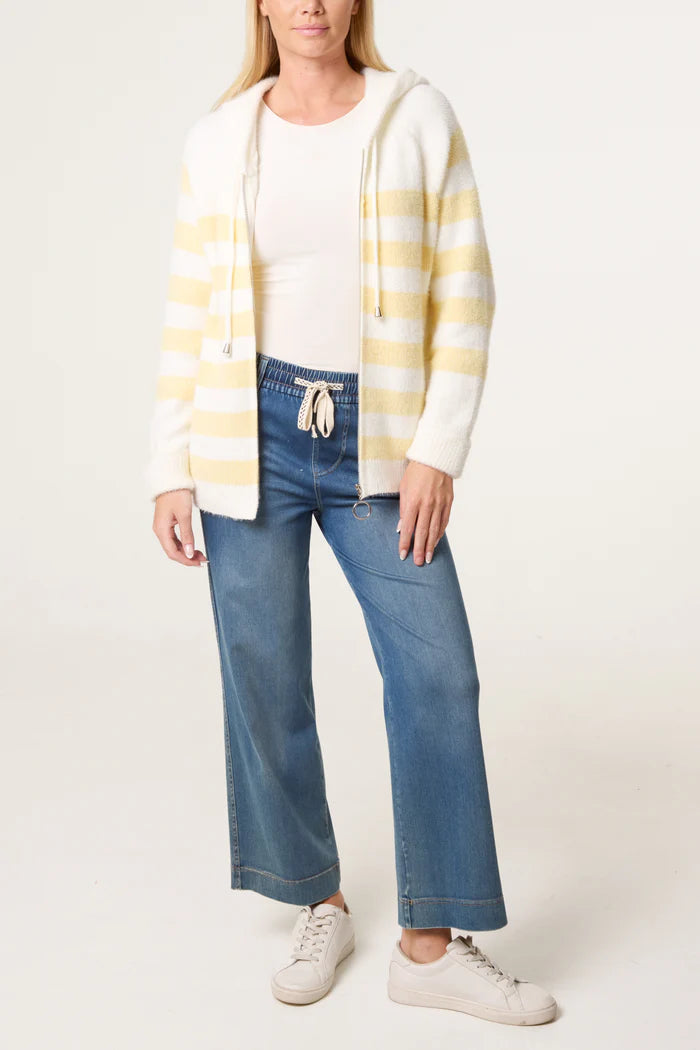 A woman wearing a zip-up cosy striped hoodie, white shirt, blue wide-leg jeans, and white trainers stands against a plain background.