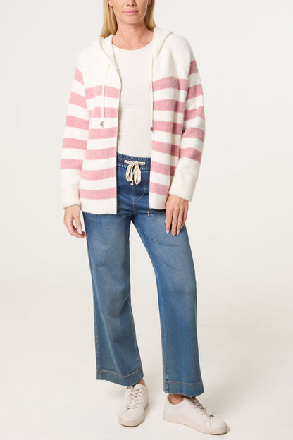 A woman stands wearing a cosy striped hoodie in white and pink, a white shirt, blue drawstring jeans, and white trainers against a plain background.