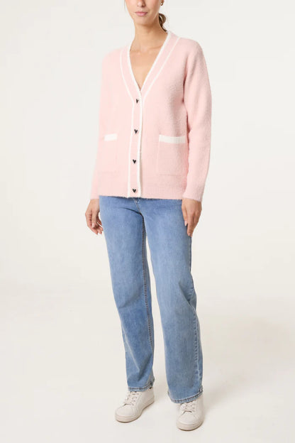 A person dressed in a light pink luxury cardigan with white trim and heart-shaped buttons, light blue jeans, and white trainers stands against a plain background.