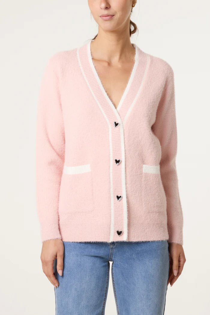 A woman wears a luxury cardigan in light pink with white edging, heart-shaped buttons, and front pockets, stylishly paired with light blue jeans.
