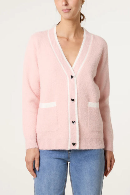 A woman wears a luxury cardigan in light pink with white edging, heart-shaped buttons, and front pockets, stylishly paired with light blue jeans.
