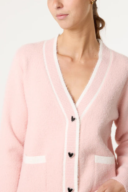 A woman wears a luxury cardigan in pale pink with white edging and black heart-shaped buttons.