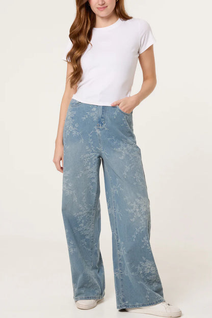 A woman in a plain white T-shirt and wide-leg, floral-print jeans made from vegan denim stands with one hand in her pocket against a neutral backdrop.