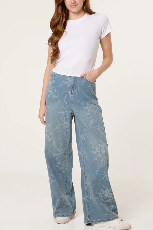 A woman in a plain white T-shirt and wide-leg, floral-print jeans made from vegan denim stands with one hand in her pocket against a neutral backdrop.