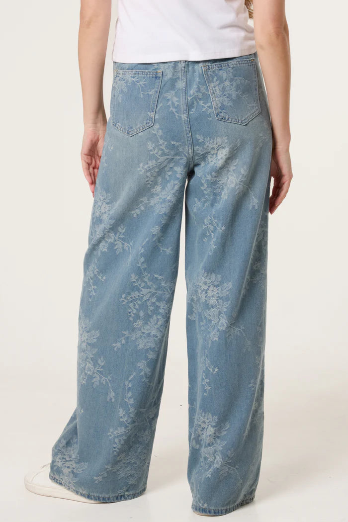 Person wearing light blue wide-leg floral print jeans featuring a subtle floral design and vintage-inspired silhouette, seen from behind, paired with a white top.