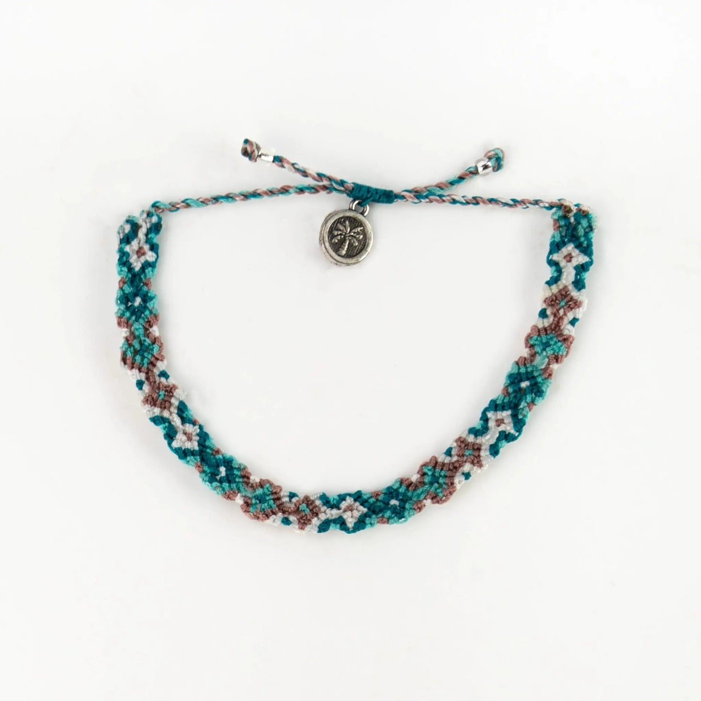 A handwoven macramé bracelet in teal, brown, and white threads, finished with a small round silver compass charm—a signature Leme Surf Bracelet inspired by Balinese jewellery.