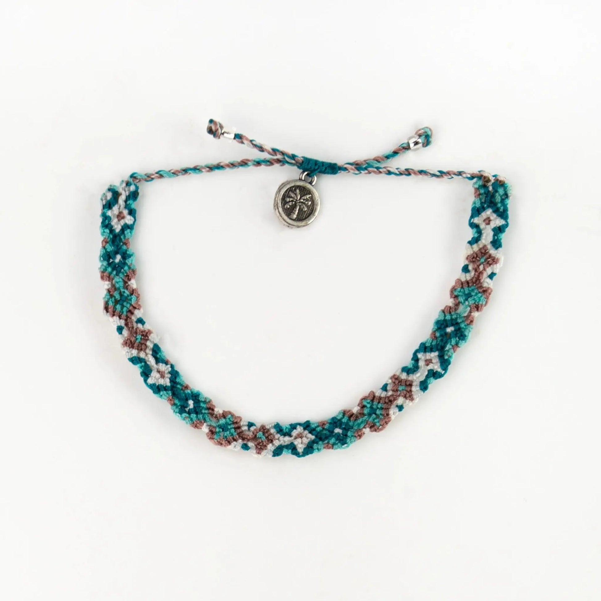 A handwoven macramé bracelet in teal, brown, and white threads, finished with a small round silver compass charm—a signature Leme Surf Bracelet inspired by Balinese jewellery.