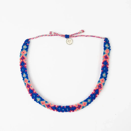 A colourful braided Leme Surf Bracelet showcasing a blue, pink, and red pattern, finished with a small round gold charm and an adjustable string fastening.