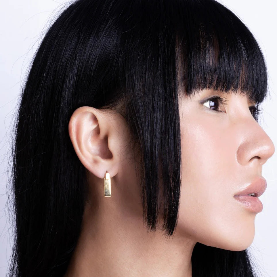 A person with straight black hair and a fringe is shown in profile, wearing a rectangular link stud earring on their left ear, set against a plain light background.