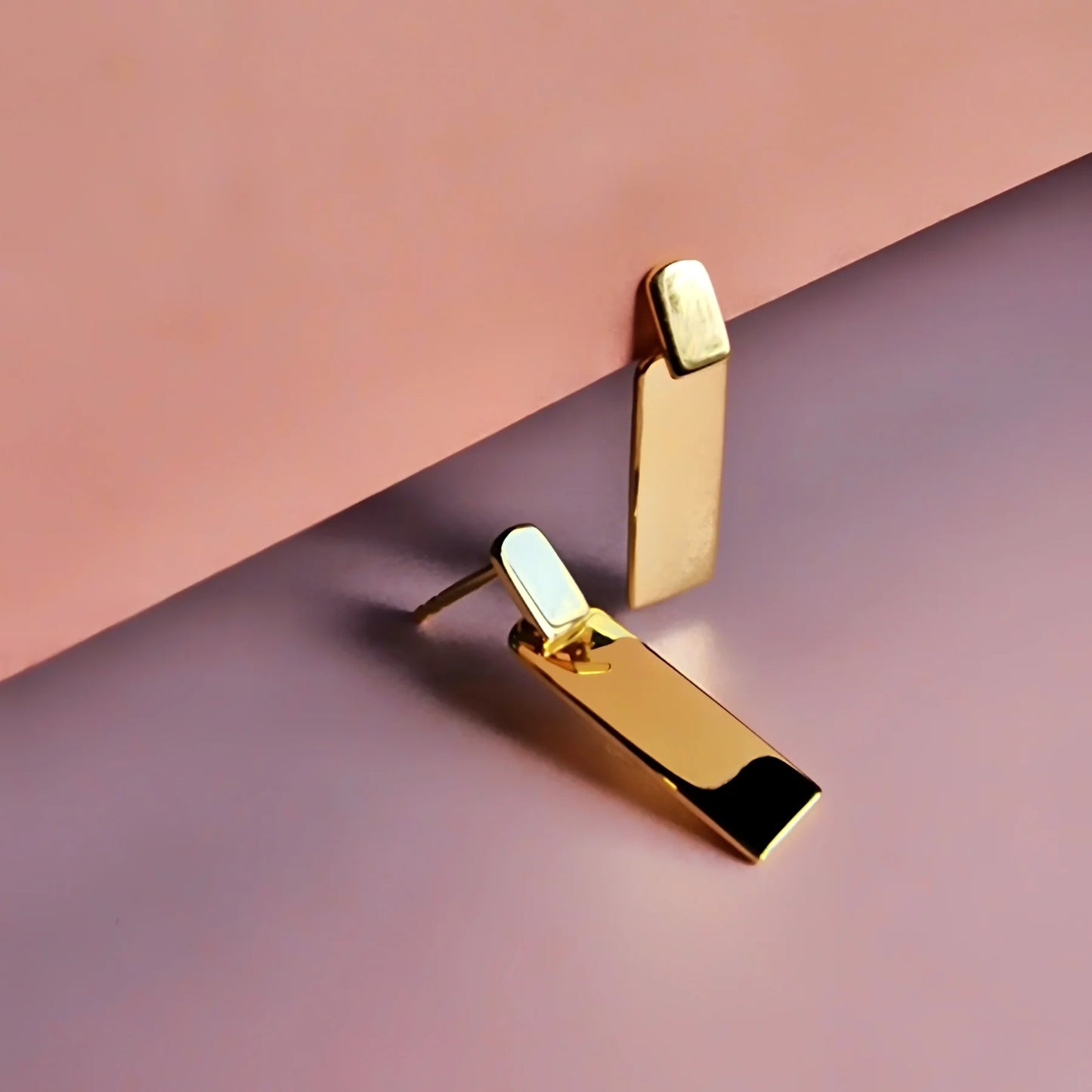 Two rectangular gold stud earrings are shown against a pink and purple background, one standing upright and the other lying flat—stylish hypoallergenic earrings ideal for any occasion.