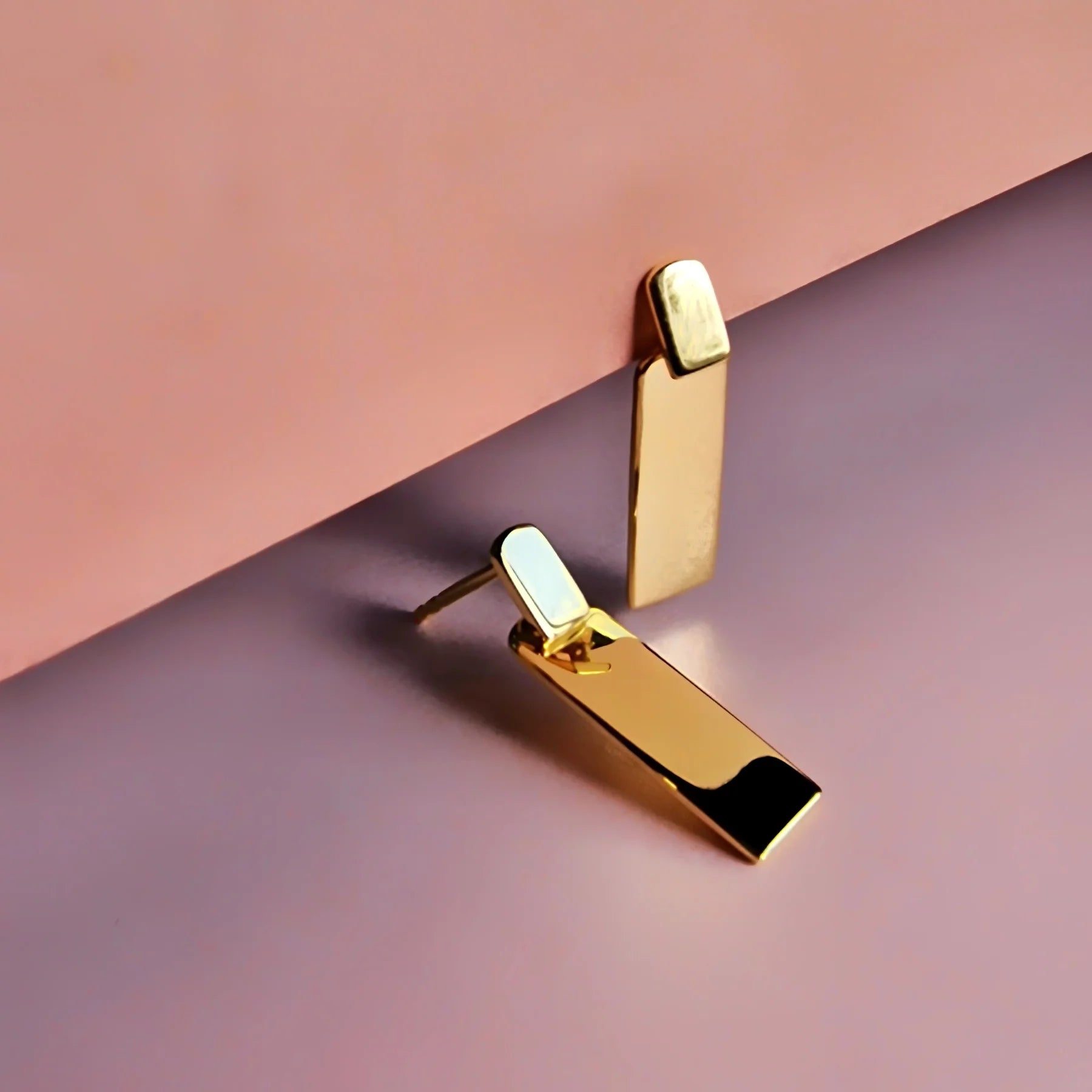 Two rectangular gold stud earrings are shown against a pink and purple background, one standing upright and the other lying flat—stylish hypoallergenic earrings ideal for any occasion.