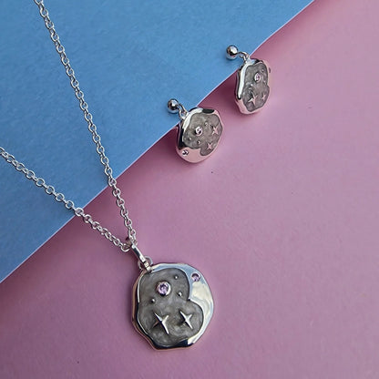 Silver necklace and celestial earrings set featuring textured circular pendants, delicate star and swirl engravings, and pink gemstones, presented on a blue and pink background.