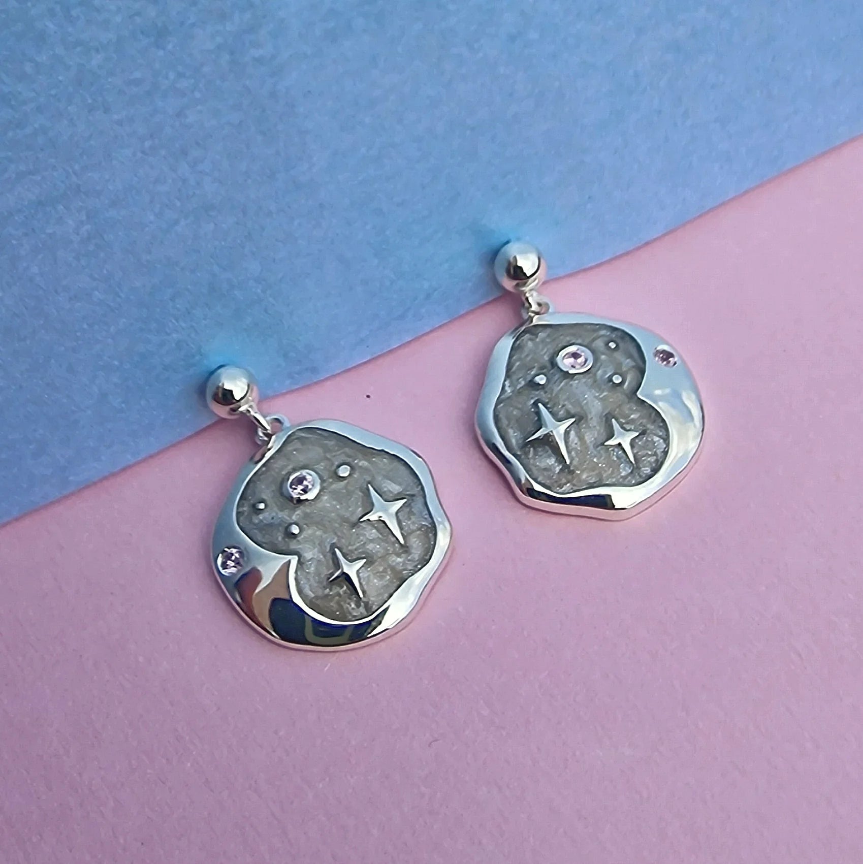 A pair of hypoallergenic silver celestial earrings with an irregular shape, adorned with star and dot designs and small gemstones, resting on a pastel pink and blue background.