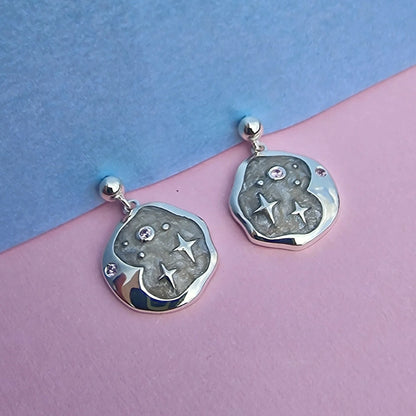 A pair of hypoallergenic silver celestial earrings with an irregular shape, adorned with star and dot designs and small gemstones, resting on a pastel pink and blue background.