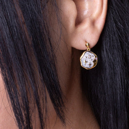 Close-up of a person's ear adorned with gold-framed celestial jewellery—an irregularly shaped earring featuring a textured, silvery-white surface and small embedded stones.