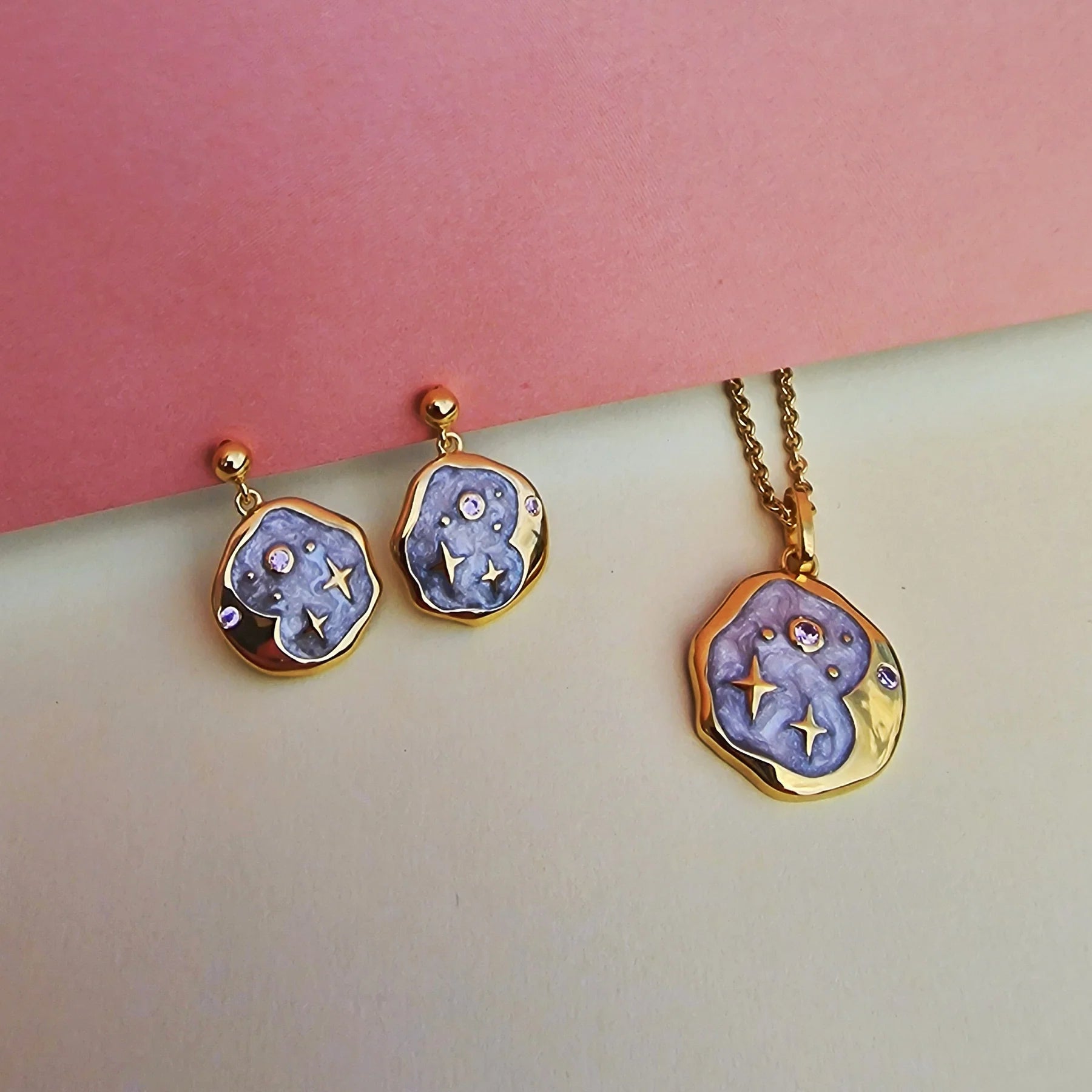 Gold-edged necklace and celestial jewellery set featuring irregular purple enamel shapes, pink opal earrings, pink stone details, and star motifs, arranged on pastel pink and beige surfaces.