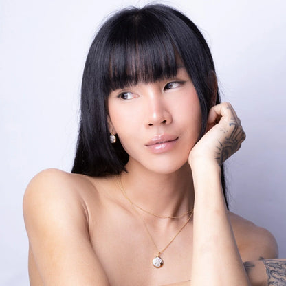Person with long black hair and fringe poses against a light background, wearing a gold celestial necklace and other gold jewellery, gently touching their face with a tattooed hand.