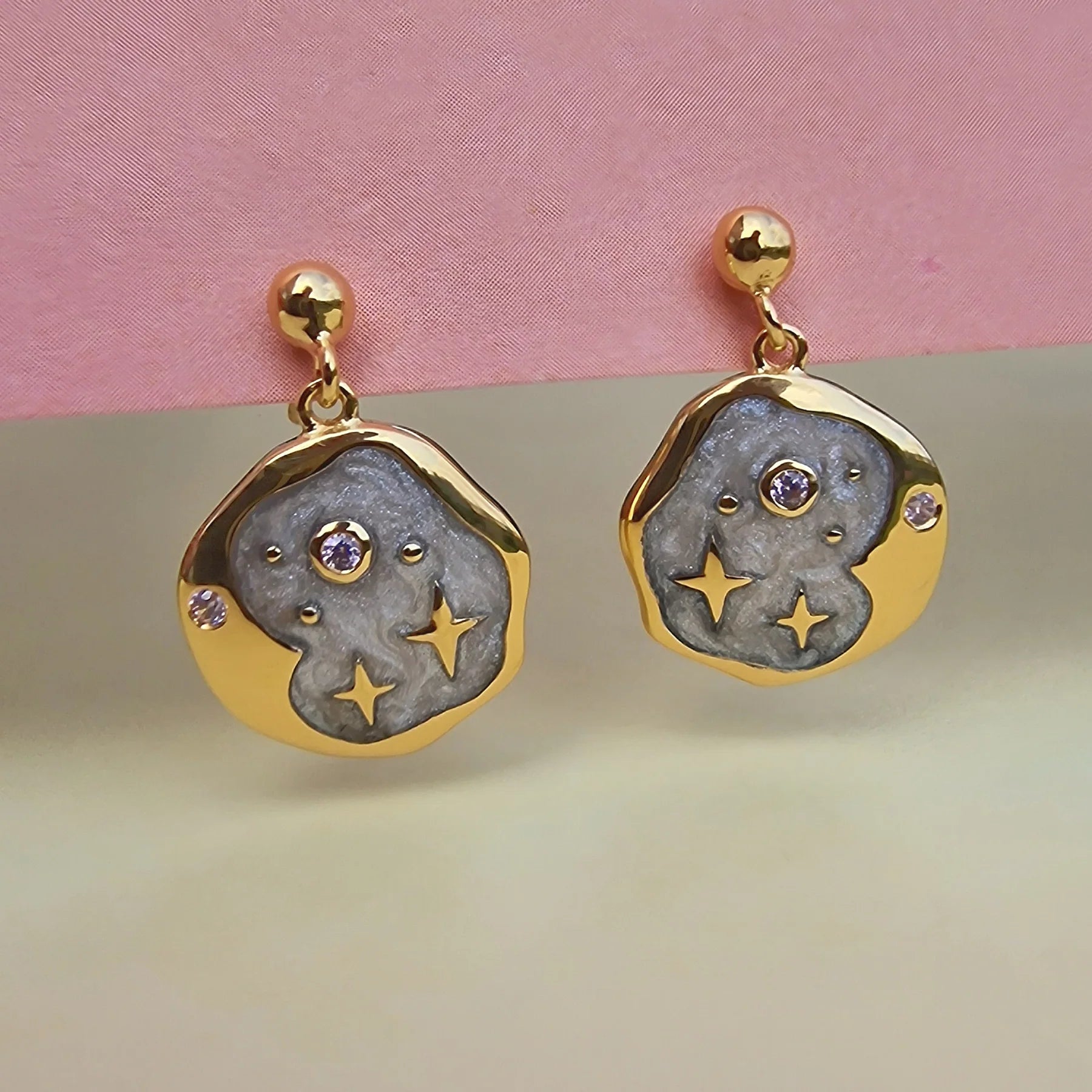 Gold and grey celestial jewellery earrings with a circular abstract design, featuring central purple stones and star-shaped cut-outs, displayed against a pink and beige background.