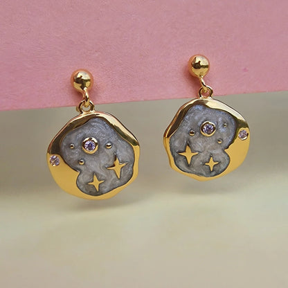 Gold and grey celestial jewellery earrings with a circular abstract design, featuring central purple stones and star-shaped cut-outs, displayed against a pink and beige background.