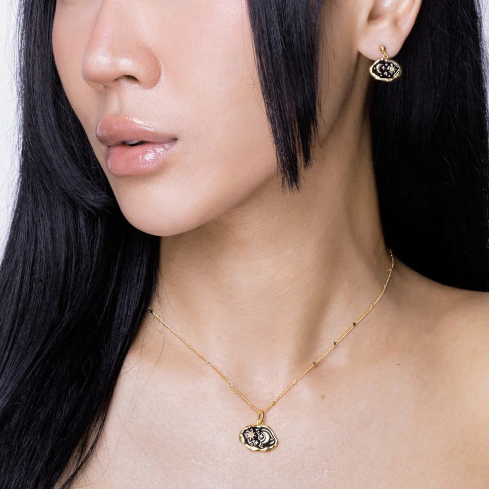 A woman with long black hair wears a gold hypoallergenic necklace and matching gold earrings featuring black and white eye-shaped pendants.