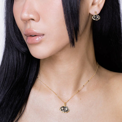 A woman with long black hair wears a gold hypoallergenic necklace and matching gold earrings featuring black and white eye-shaped pendants.