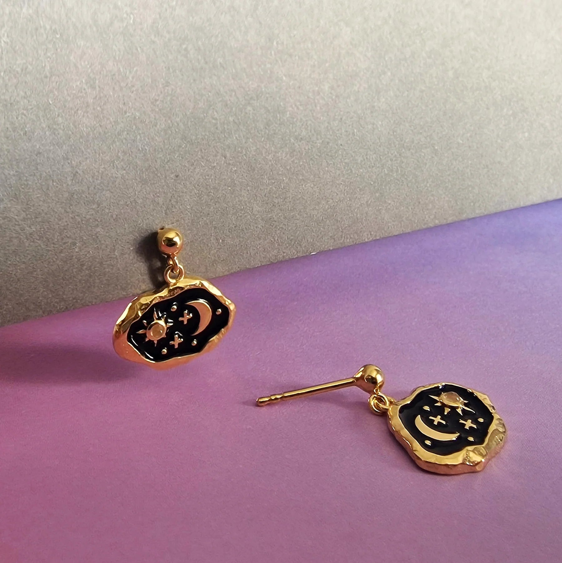 A pair of gold hypoallergenic and black earrings featuring celestial designs, including a moon and stars, displayed against a purple and grey background.