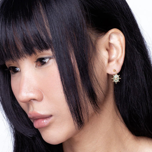 A woman with straight black hair wears a hypoallergenic gold-plated geometric stud earring with a light blue centre on her left ear, facing left against a plain white background.