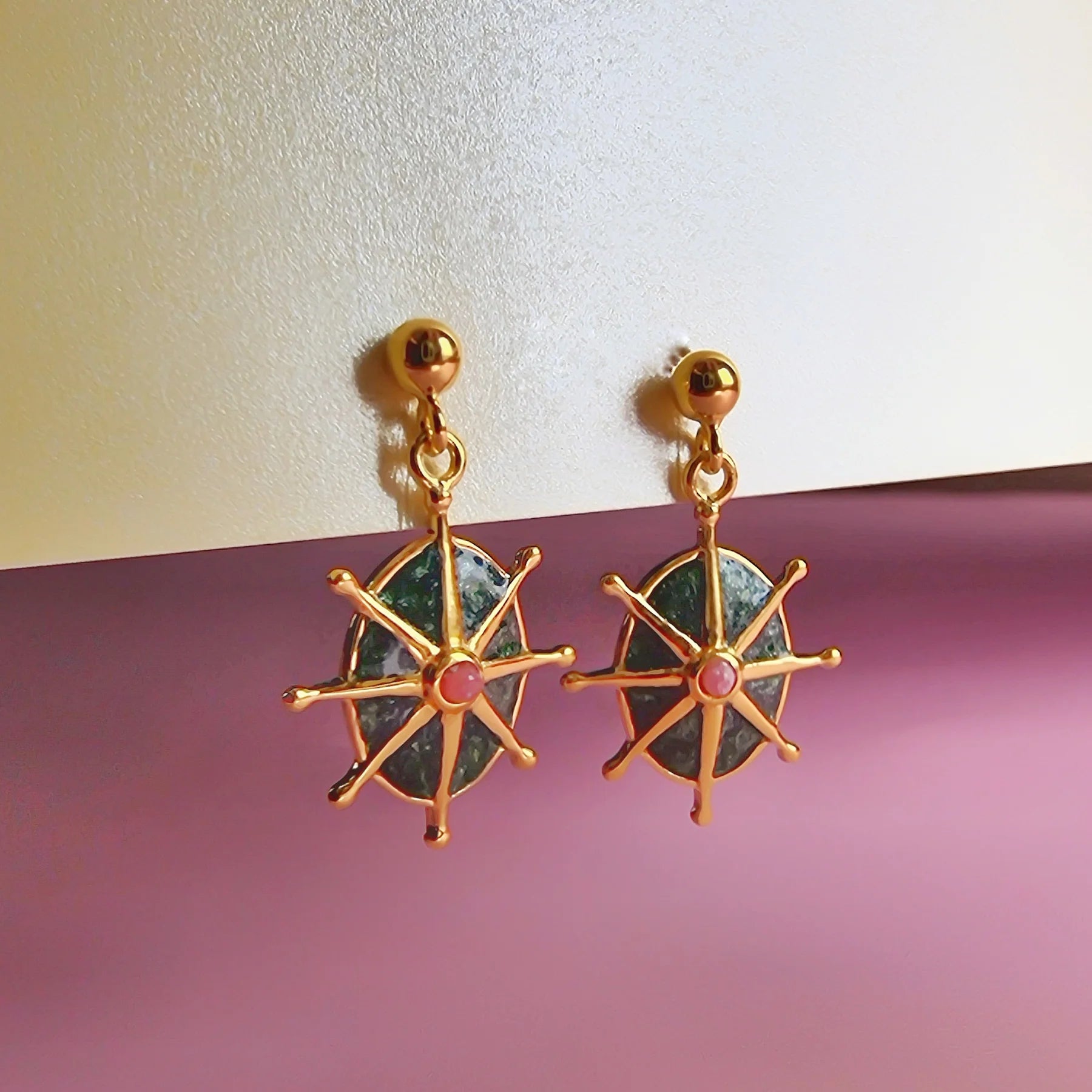 Gold-plated brass earrings with ship’s wheel designs, each featuring a central emerald green star stud and small pink accents, set against a pale and purple background.