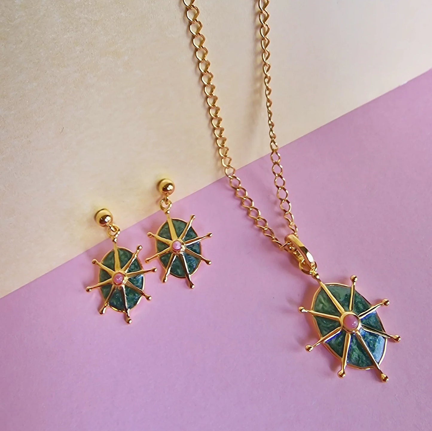 Gold necklace and earrings set featuring green enamel pendants and pink geometric accents, displayed against a two-tone beige and pink backdrop.