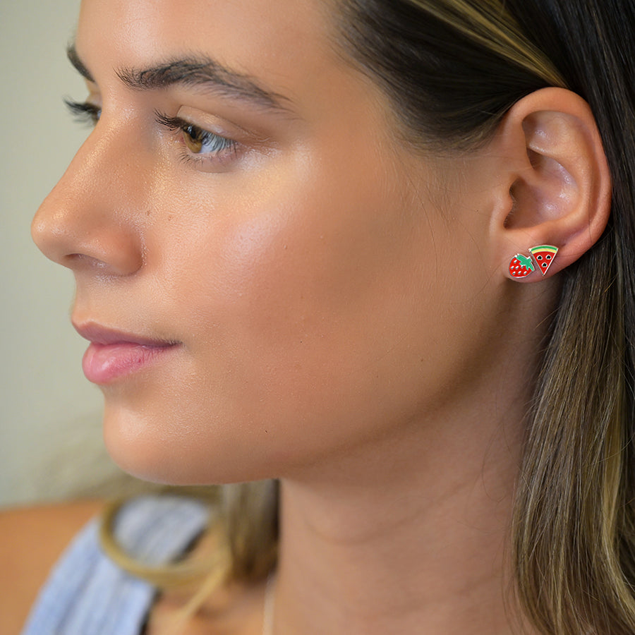 An individual wears sterling silver earrings featuring vibrant fruit themes: one a strawberry, the other a slice of watermelon. With red and green enamel adding flair, their expression remains neutral against the light background.