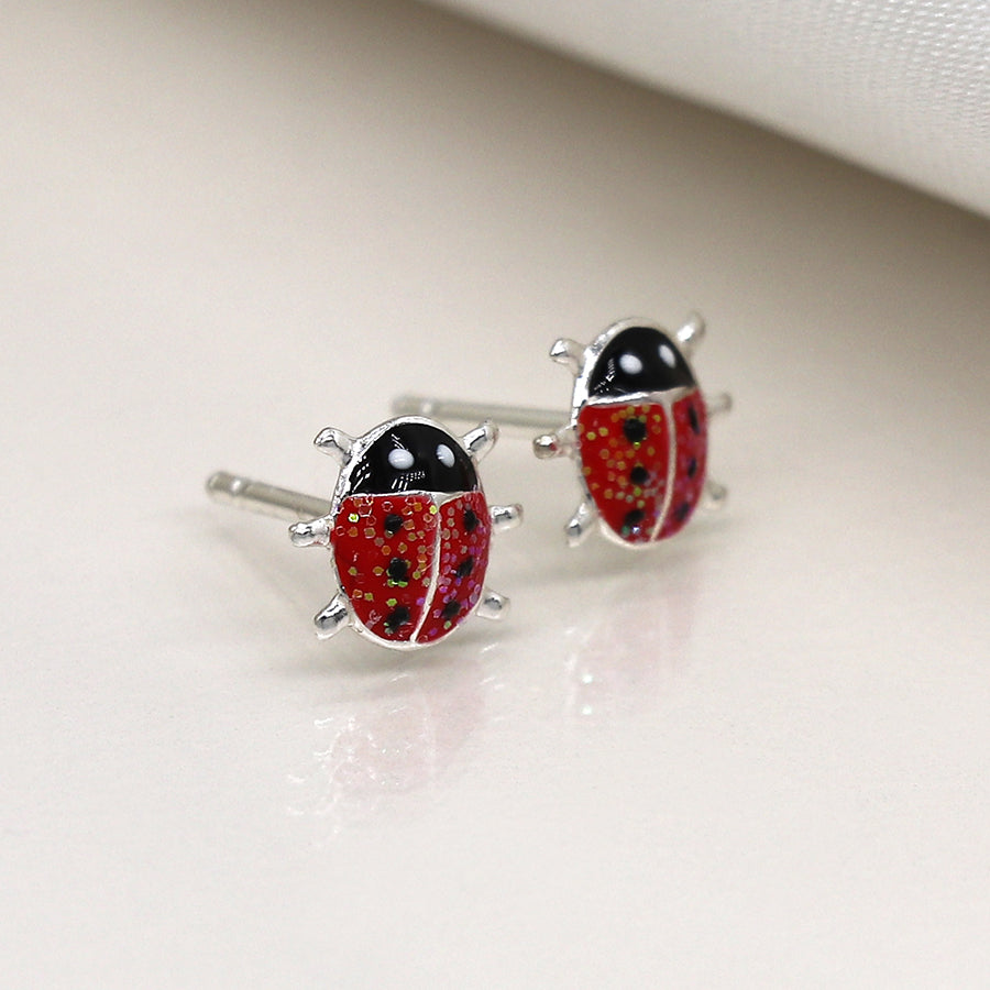 Close-up of handcrafted enamel ladybird earrings with red and black details, featuring small sterling silver legs, resting on a light-coloured surface.
