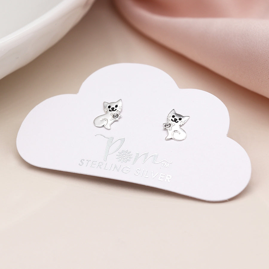 Two small sterling silver stud earrings, featuring a charming cat design, are displayed on a cloud-shaped card against a soft pink fabric background.