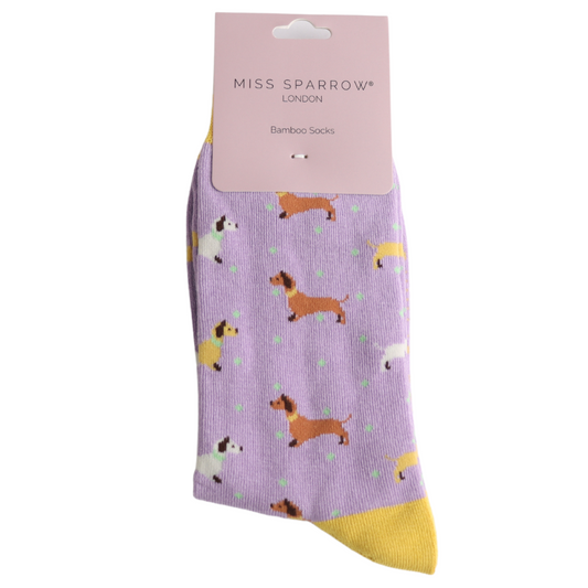 A single lilac bamboo sock featuring a pattern of various dogs, including dachshunds, with a yellow toe and a pink Miss Sparrow London vegan socks label attached at the top.