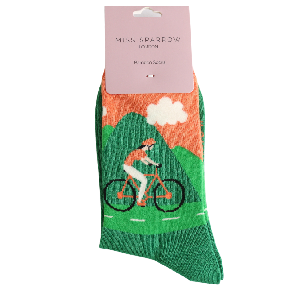Green bamboo socks featuring a cyclist on a mountain path, crafted from sustainable bamboo fibres and packaged with a pink Miss Sparrow London Bamboo Socks label.
