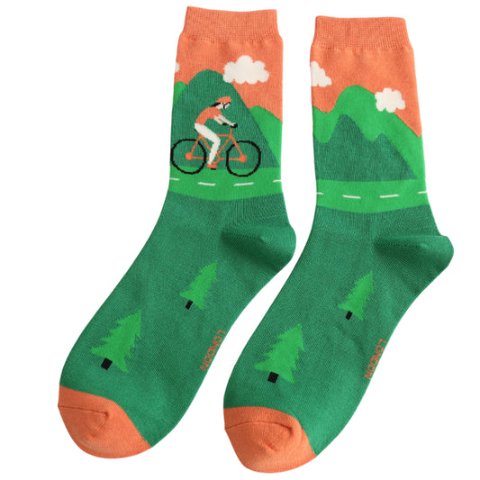 Pair of green and orange socks featuring a person cycling on a road with mountains, trees, and clouds—crafted from sustainable bamboo fibres for a vegan-friendly design.