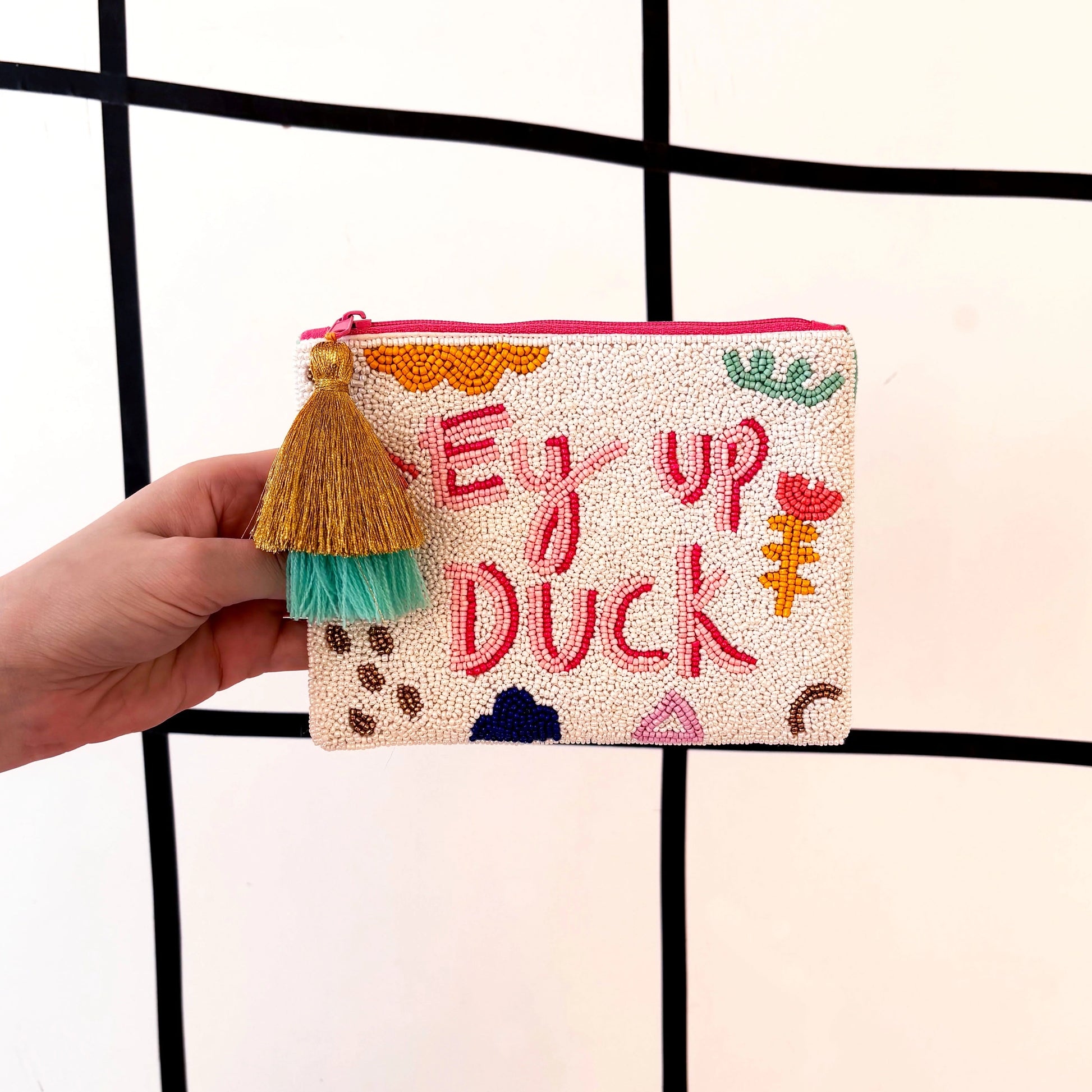 A hand holds an Ey Up Duck pouch with a tassel, showcasing colourful embroidery of British slang on black grid lines—an ideal gift for lovers of British sayings.