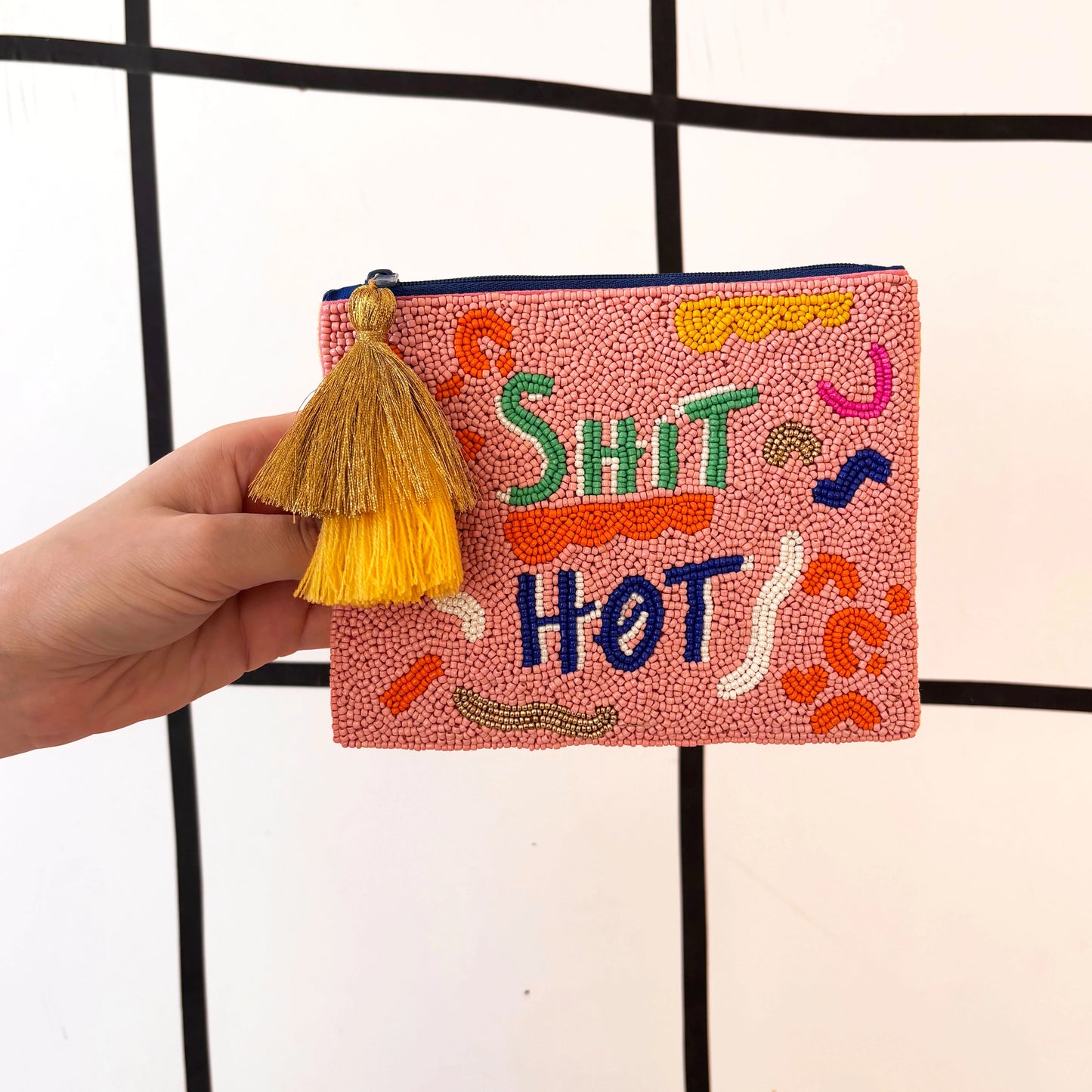 A hand holds a pink hand-beaded pouch with yellow tassels, colourful abstract designs, and the words SHIT HOT embroidered on the front, featuring intricate glass bead detailing.