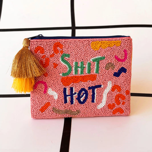 A pink hand-beaded pouch featuring colourful text that reads SHIT HOT, adorned with abstract patterns and finished with a gold and yellow velvet tassel on the zip.