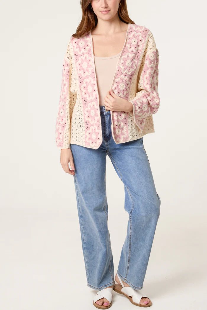 A woman wearing a light pink and cream floral crochet cardigan, a beige top, blue jeans, and white sandals stands against a plain backdrop.
