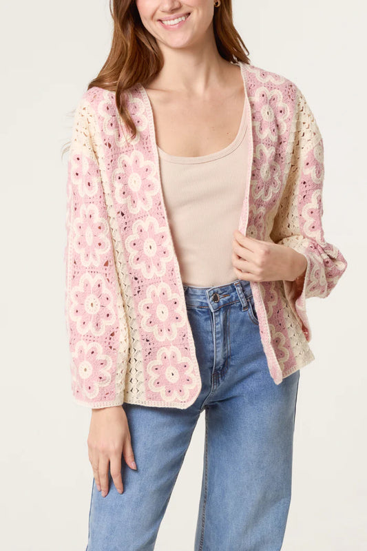 Woman wearing a cream and pink floral crochet cardigan with vintage appeal, a beige vest top, and blue jeans, standing against a plain background.
