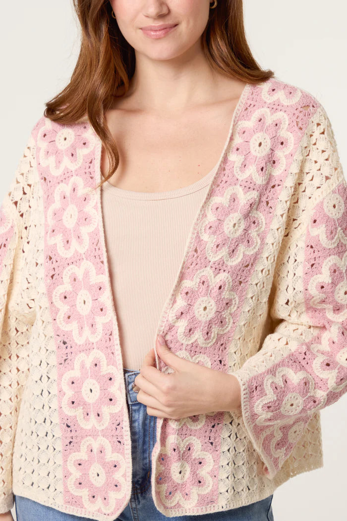 A woman wears a cream floral crochet cardigan with pink and white patterns, layered over a beige top and blue jeans, evoking vintage charm.